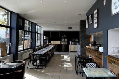 Salle lumineuse avec grandes baies vitrées, tables en bois et vue sur une rue calme bordée de bâtiments.
