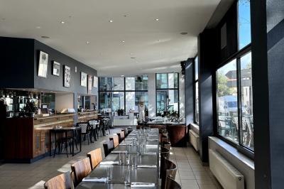 Salle lumineuse avec grandes baies vitrées, tables en bois et vue sur une rue calme bordée de bâtiments.