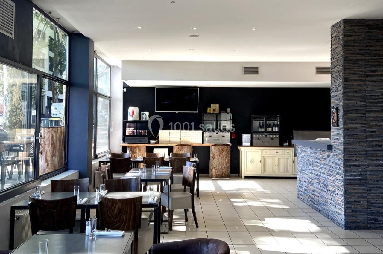 Salle de restaurant lumineuse avec tables dressées, grandes baies vitrées et comptoir en pierre au fond.