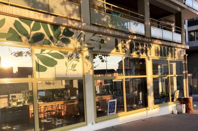 Salle lumineuse avec grandes baies vitrées, tables en bois et vue sur une rue calme bordée de bâtiments.