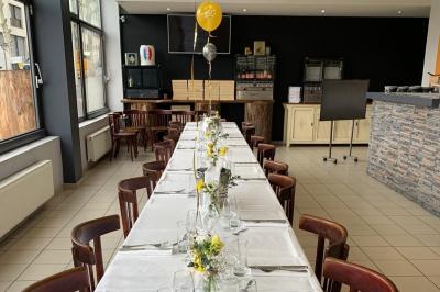 Salle lumineuse avec grandes baies vitrées, tables en bois et vue sur une rue calme bordée de bâtiments.