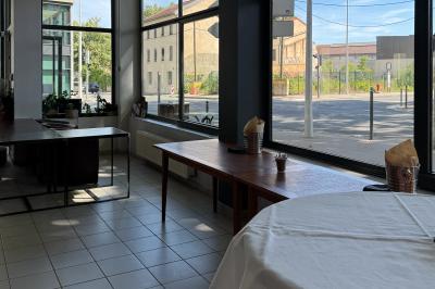 Salle lumineuse avec grandes baies vitrées, tables en bois et vue sur une rue calme bordée de bâtiments.