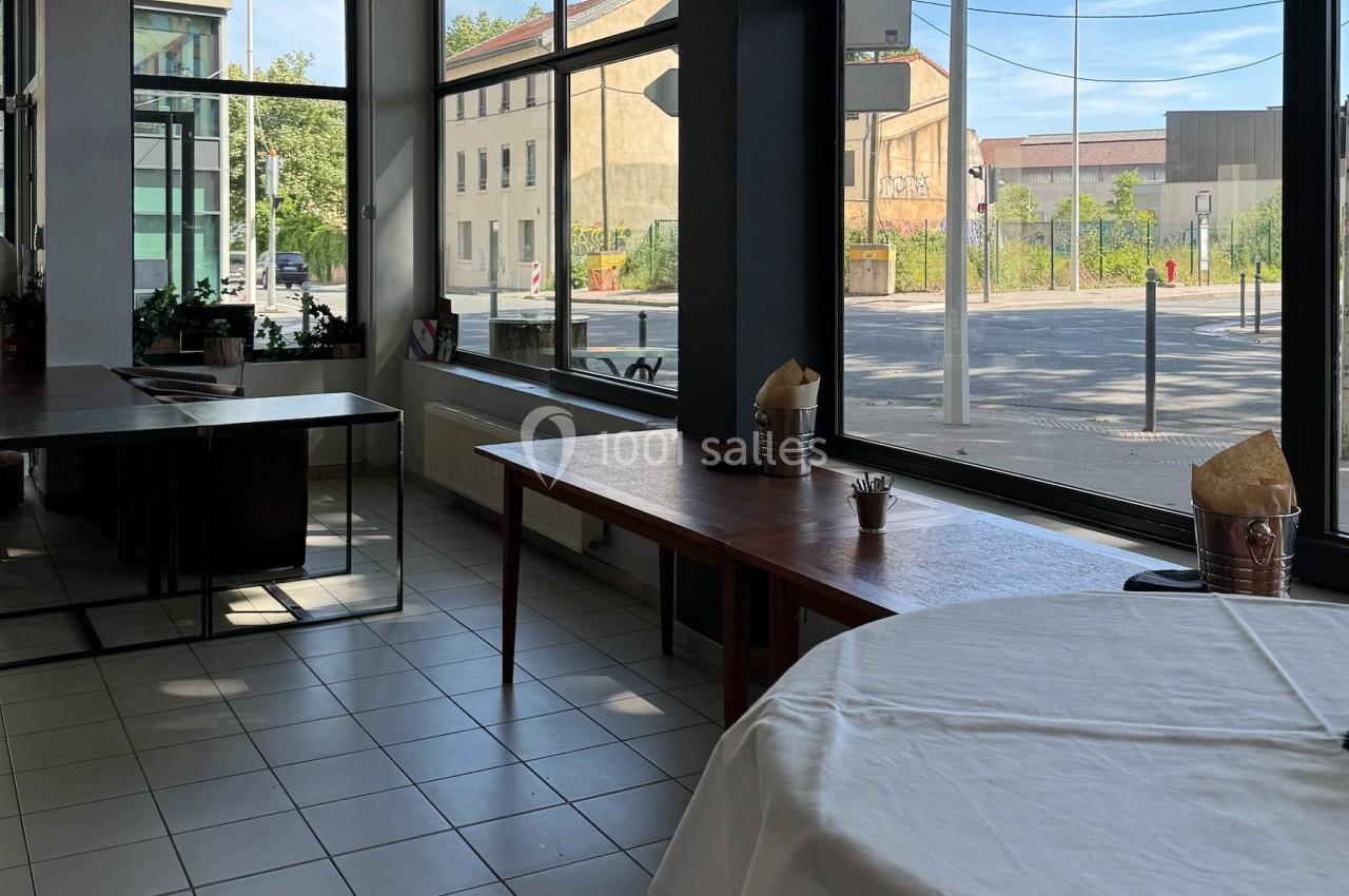 Salle lumineuse avec grandes baies vitrées, tables en bois et vue sur une rue calme bordée de bâtiments.
