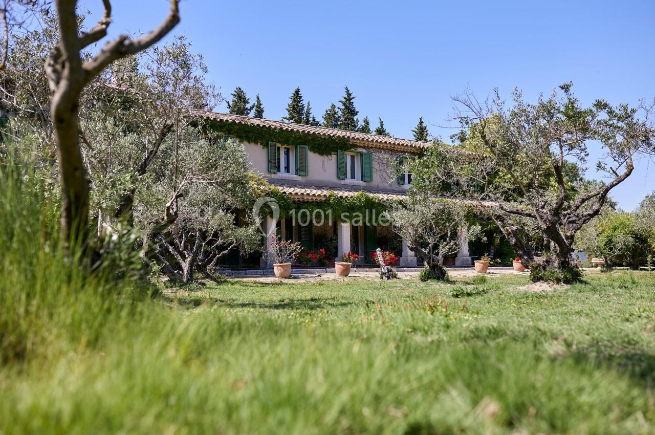 Maison provençale entourée d'oliviers et de pots de fleurs, avec un jardin verdoyant sous un ciel bleu.
