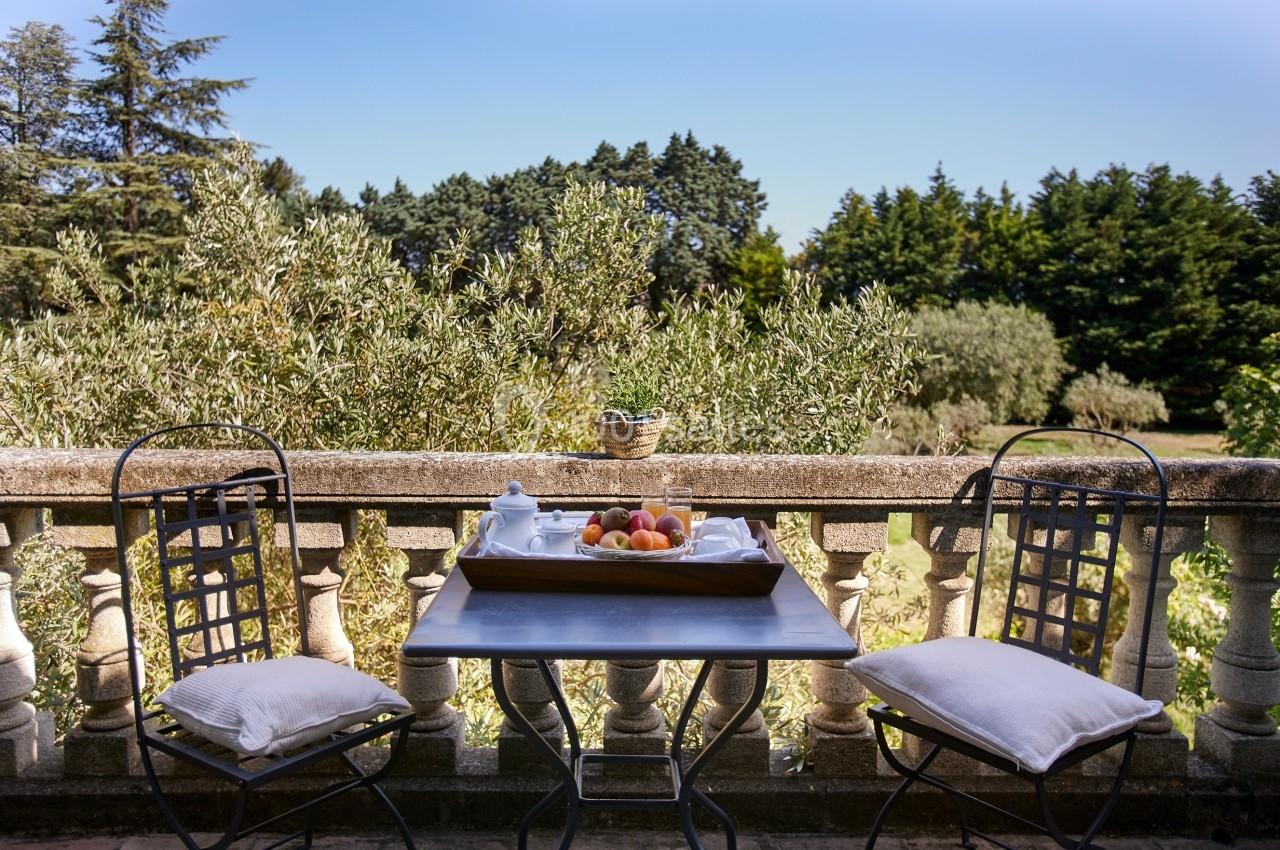 Table en fer forgé avec coussins, plateau de fruits et théière sur une terrasse surplombant un jardin arboré.