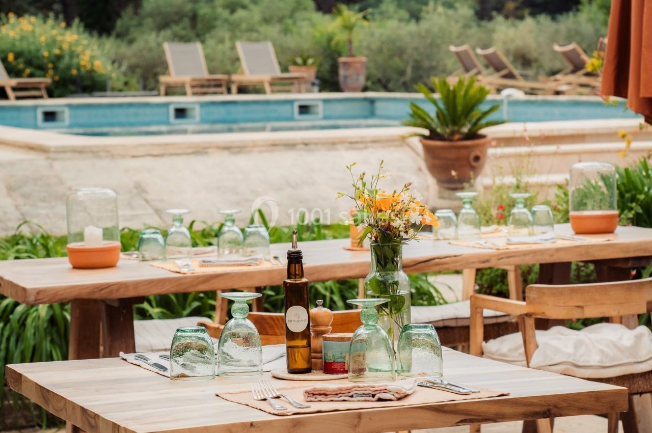 Tables en bois dressées en extérieur près d'une piscine, entourées de verdure et de chaises longues.