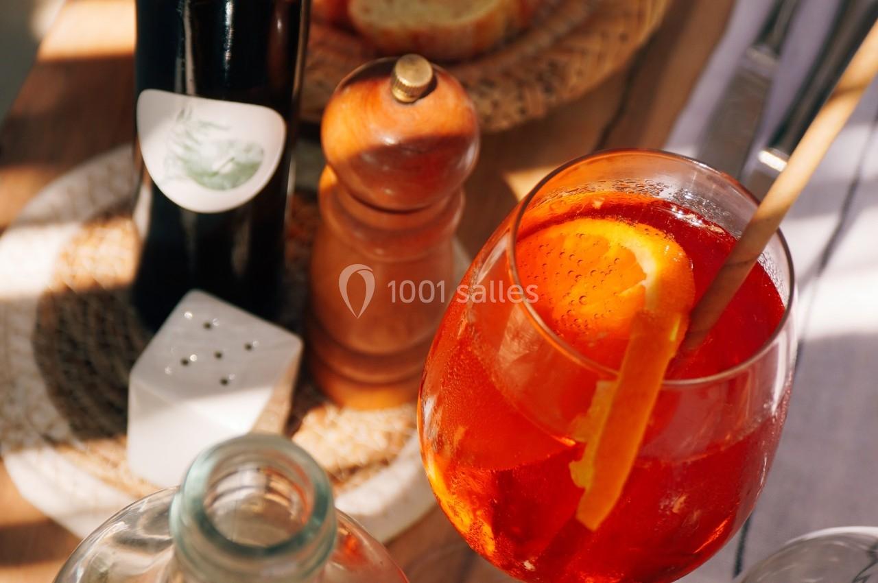 Verre de cocktail orange avec une tranche d'orange, entouré de pain, huile, poivrier et carafe sur une table en bois.