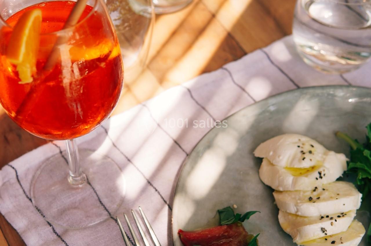 Assiette avec mozzarella, tomates et basilic, accompagnée d'un verre de cocktail orange sur une table ensoleillée.