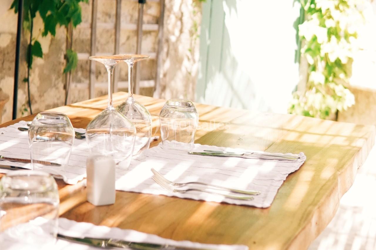 Table en bois dressée avec des verres et des couverts, située en extérieur près d'un mur couvert de plantes grimpantes.