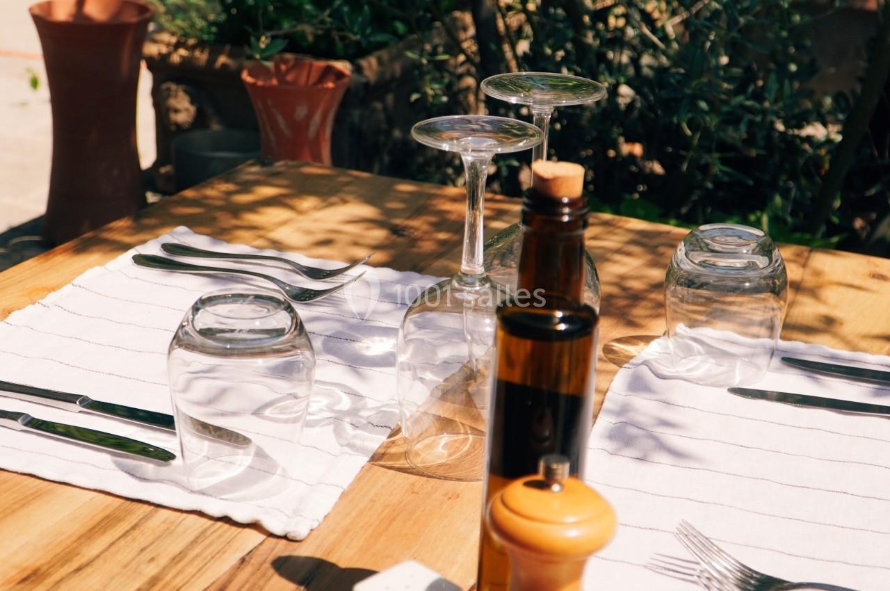 Table en bois dressée avec couverts, verres, huile, vinaigre, sel et poivre, entourée de plantes en extérieur.