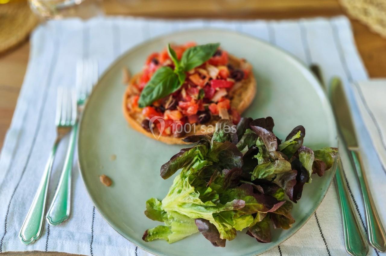 Tartine garnie de tomates, olives et basilic, accompagnée de salade verte sur une assiette posée sur une nappe rayée.
