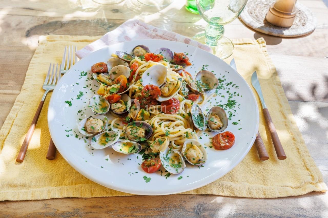 Assiette de spaghetti aux palourdes, tomates cerises et persil, servie sur une table ensoleillée avec des couverts.