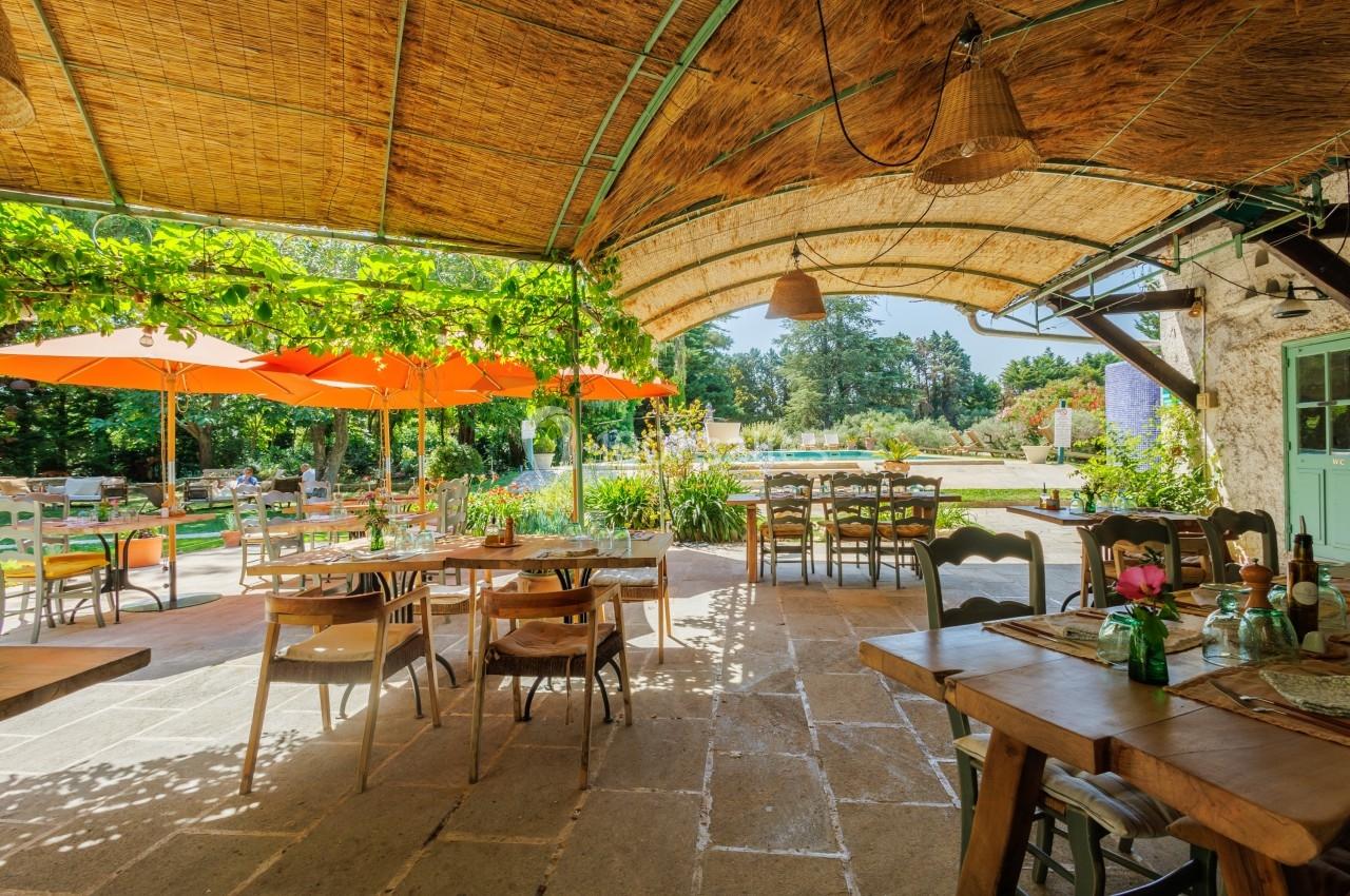 Terrasse ombragée avec tables en bois, vue sur un jardin verdoyant et parasols orange en arrière-plan.