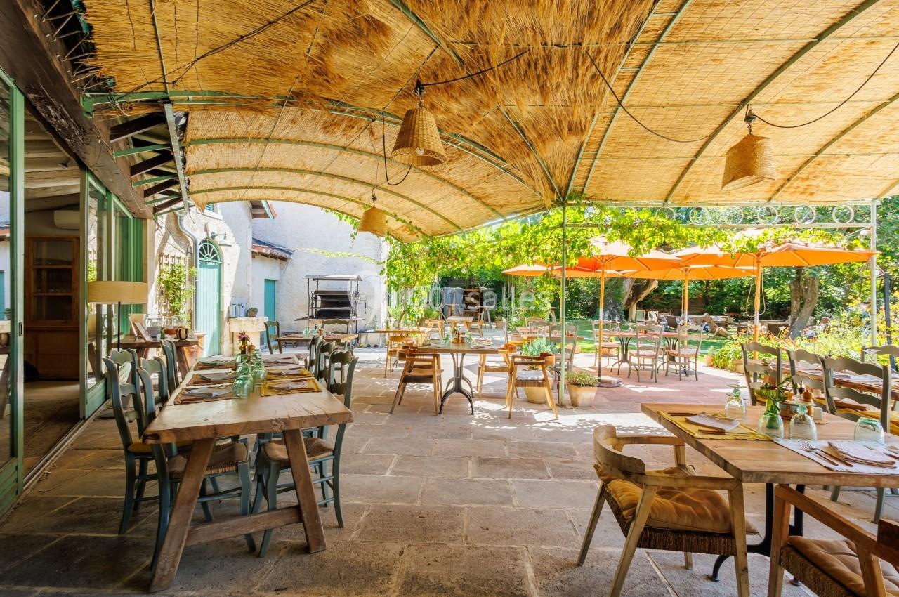 Terrasse d'un restaurant avec tables en bois, chaises rustiques, toit en canisse et parasols orange dans un cadre verdoyant.