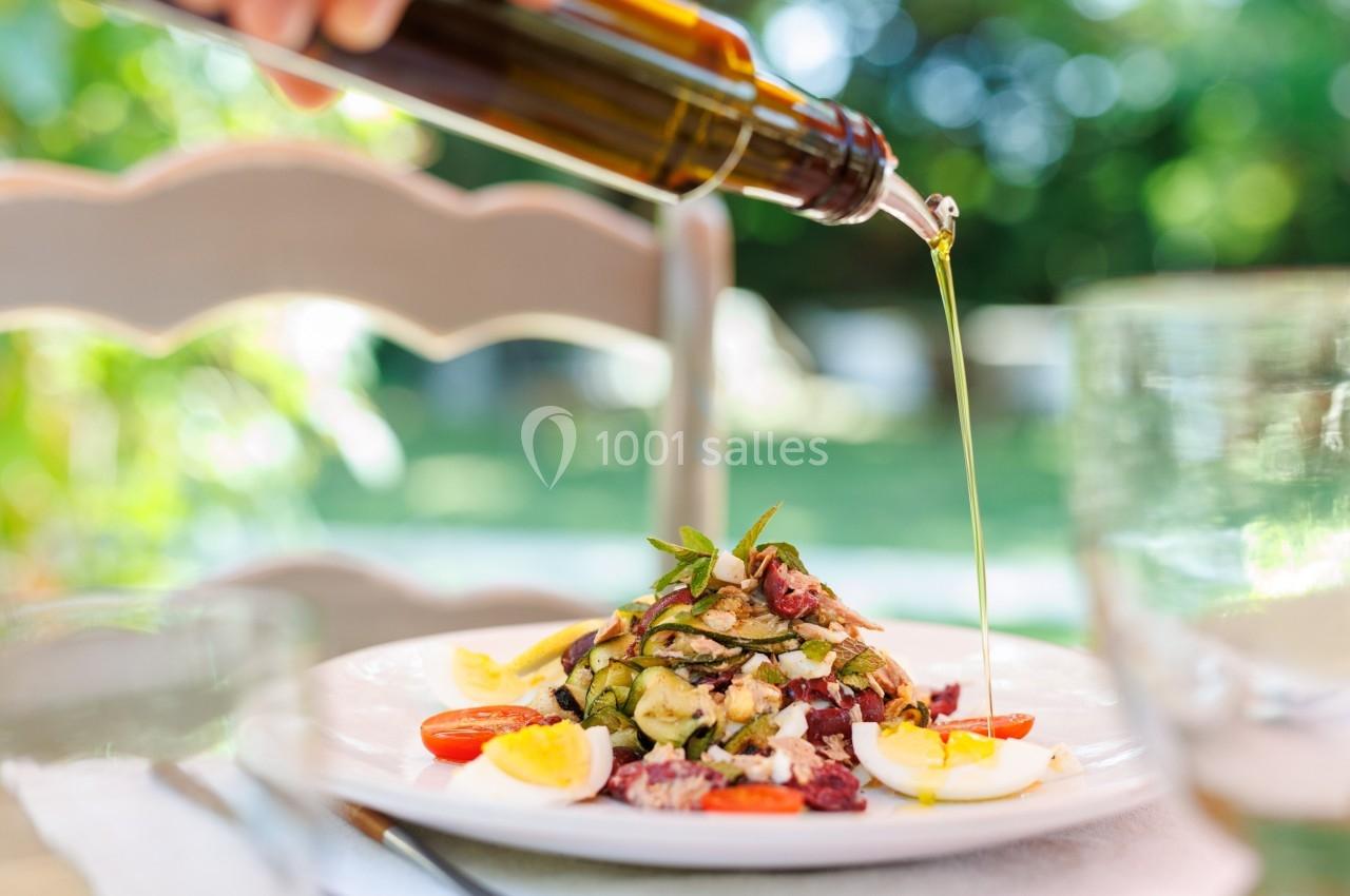 Assiette de salade garnie de légumes, œufs et tomates, arrosée d'huile d'olive versée depuis une bouteille.