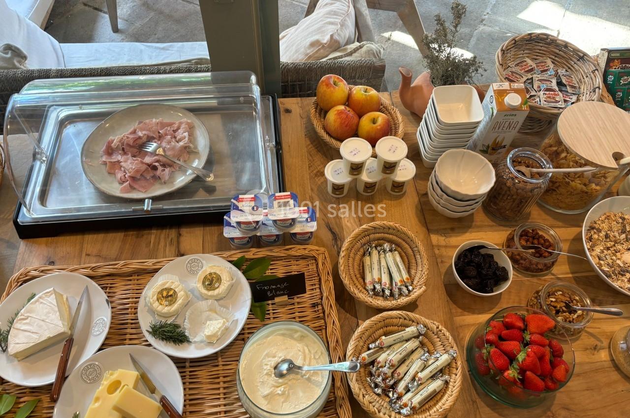 Buffet de petit-déjeuner avec charcuterie, fromages, fruits, céréales, yaourts et confitures sur une table en bois.