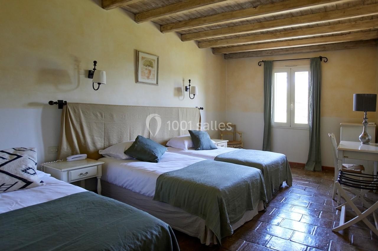 Chambre lumineuse avec trois lits simples, murs clairs, plafond en poutres apparentes et fenêtre avec rideaux verts.