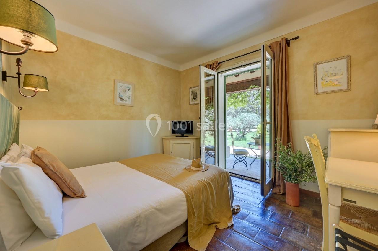 Chambre lumineuse avec lit simple, bureau, télévision et porte-fenêtre donnant sur un jardin verdoyant.