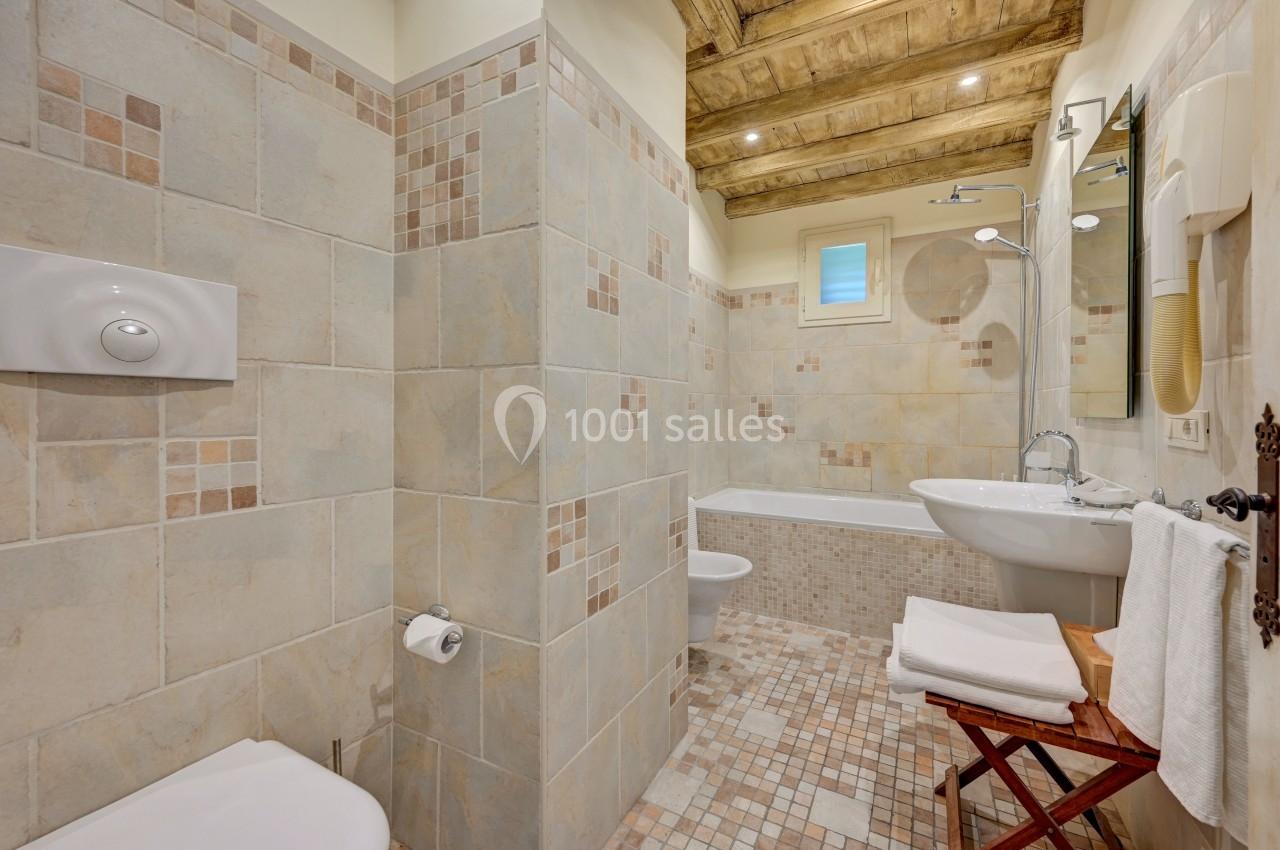 Salle de bain avec carrelage beige, baignoire, lavabo, toilettes et plafond en bois apparent.
