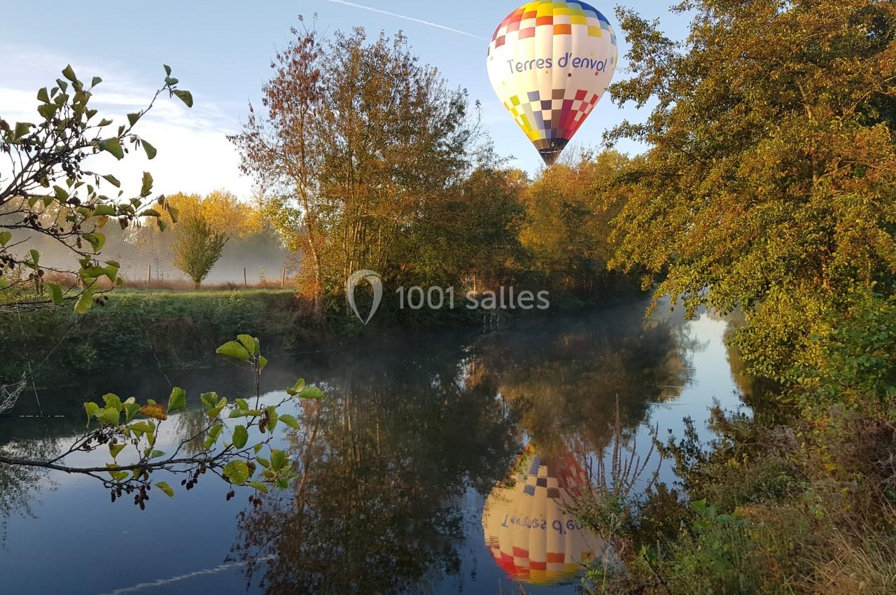 Montgolfière colorée survolant un paysage automnal avec rivière calme et arbres reflétés dans l'eau.