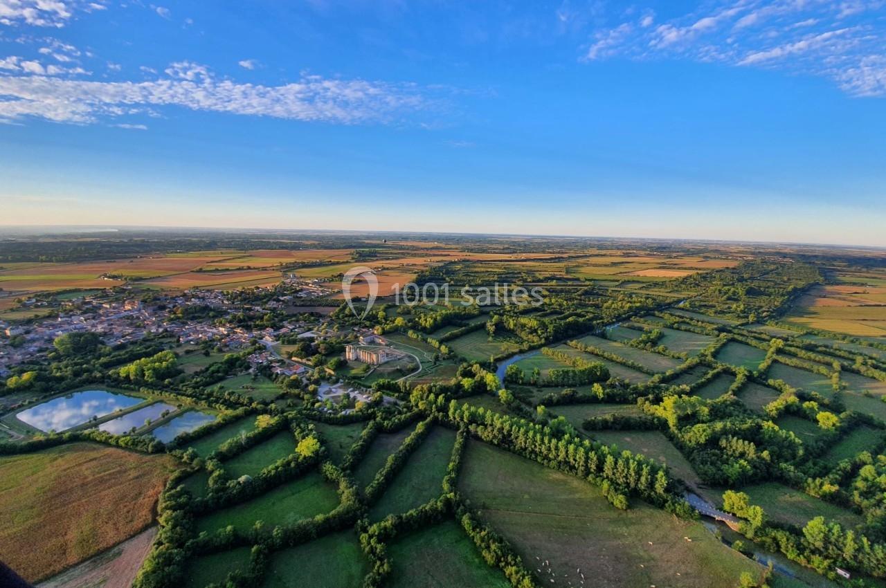 Vue aérienne d'une campagne verdoyante avec champs, haies, étangs et un village sous un ciel bleu dégagé.