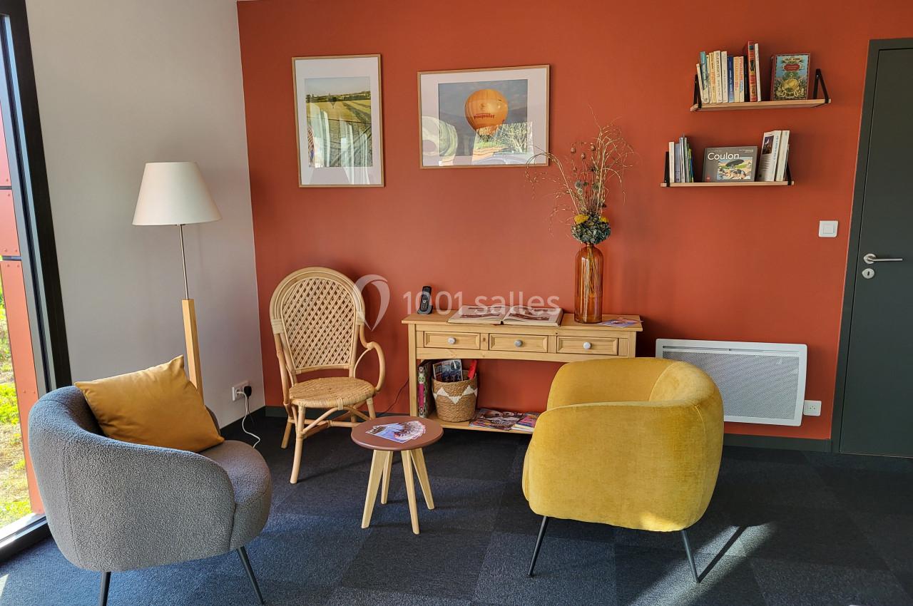Coin salon avec deux fauteuils, une chaise en rotin, une table basse, un bureau en bois et un mur orange décoré.