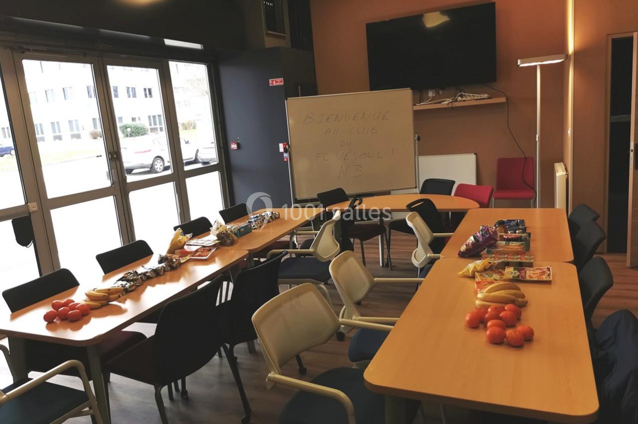 Salle de réunion avec tables disposées en U, garnies de fruits et collations, tableau blanc et chaises autour.