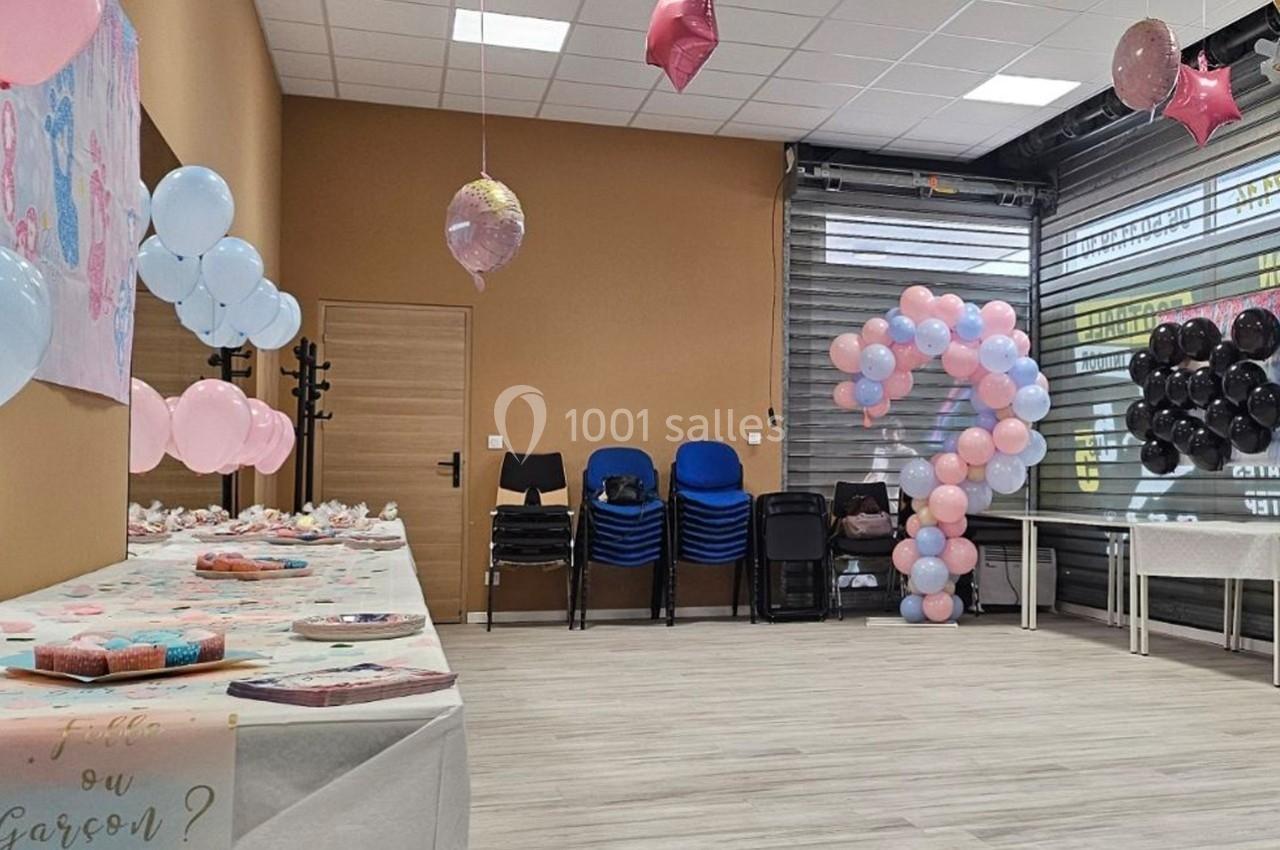 Salle décorée pour une fête avec des ballons roses, bleus et noirs, tables dressées et chaises empilées au fond.