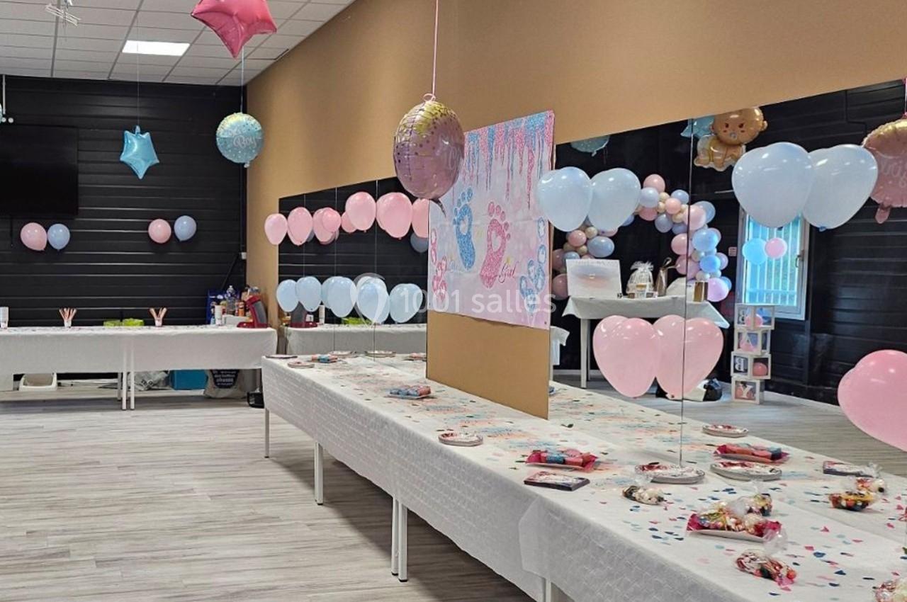 Salle décorée pour une fête avec ballons roses et bleus, tables dressées avec friandises et une affiche centrale.