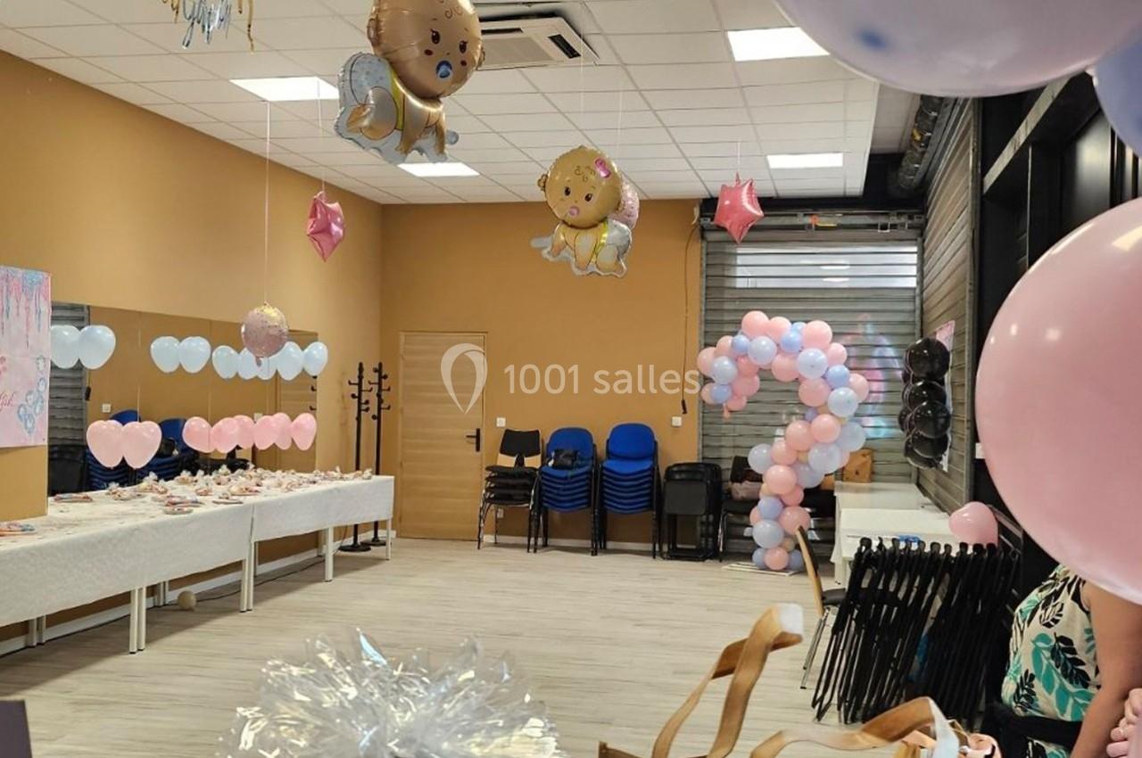 Salle décorée pour une fête avec ballons roses et bleus, tables dressées et chaises empilées au fond.