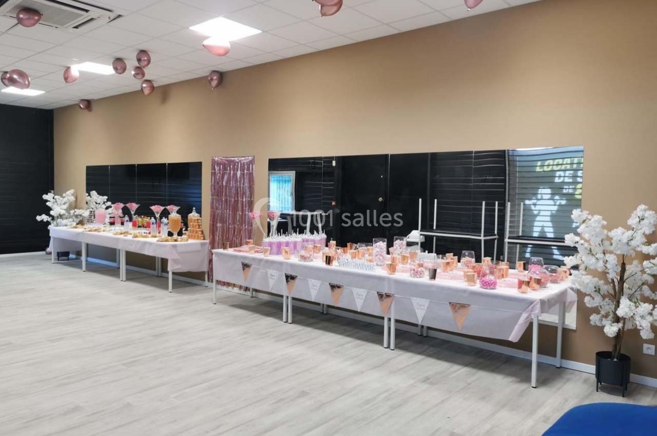 Tables décorées pour une réception avec nappes blanches, décorations roses et arbres artificiels blancs dans une salle.