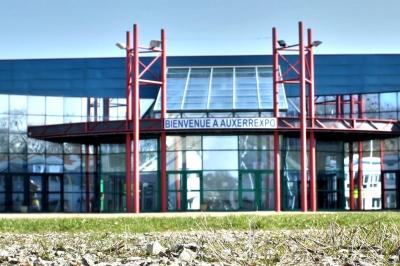Façade illuminée du bâtiment Auxerrexpo de nuit, avec des visiteurs entrant et deux tracteurs exposés devant.