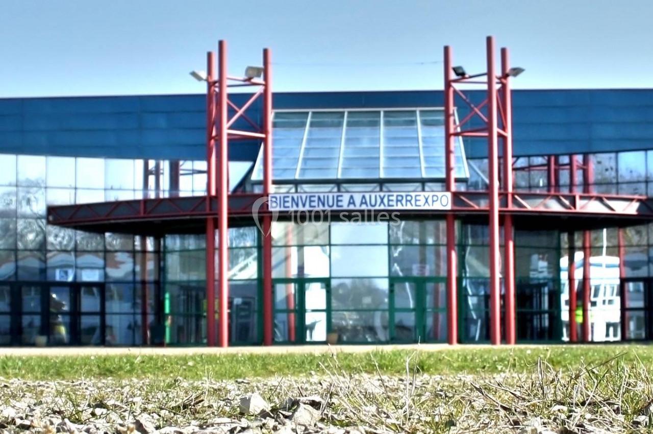 Façade vitrée d'un bâtiment moderne avec une entrée centrale et des structures rouges, vue depuis un espace extérieur.