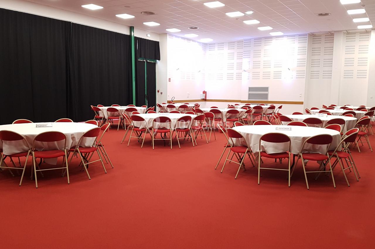 Salle de réception avec tables rondes dressées de nappes blanches et chaises rouges, sol en moquette rouge.