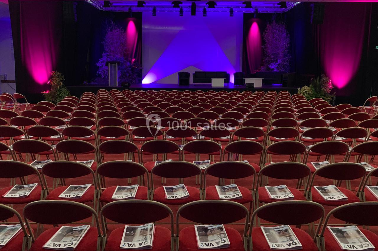 Salle de conférence avec des rangées de chaises rouges, des brochures posées sur chaque siège et une scène éclairée en…