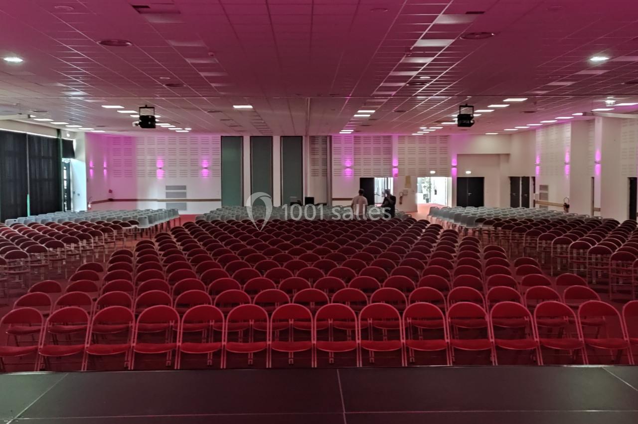 Salle de conférence avec rangées de chaises rouges alignées, éclairage rose et scène au premier plan.