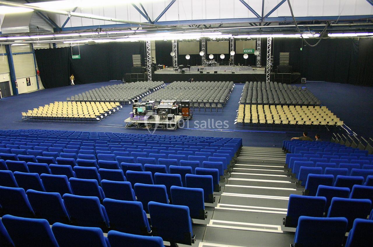Salle de spectacle vide avec sièges bleus et beiges, scène équipée de projecteurs et matériel technique au centre.