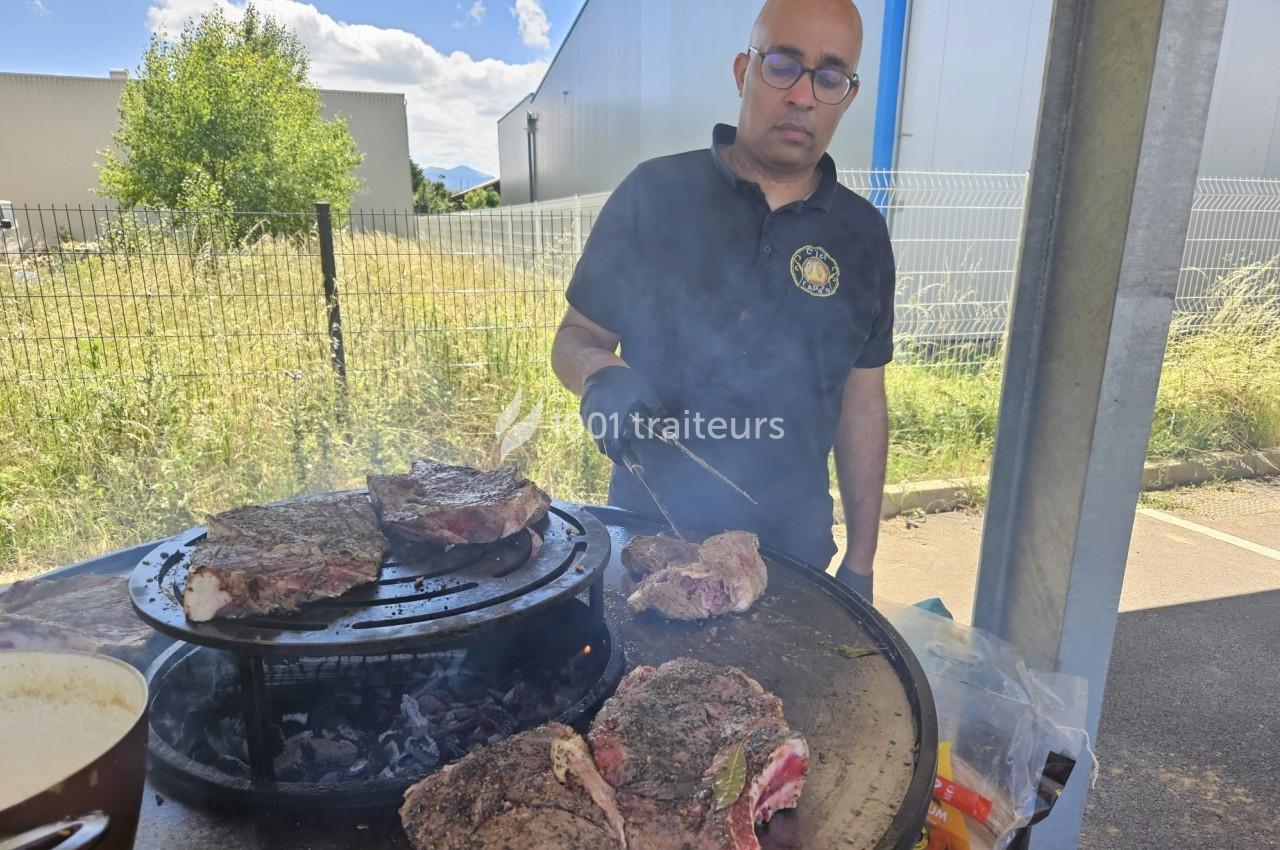 Un homme fait griller de la viande sur un barbecue circulaire en extérieur, entouré de bâtiments et de végétation.