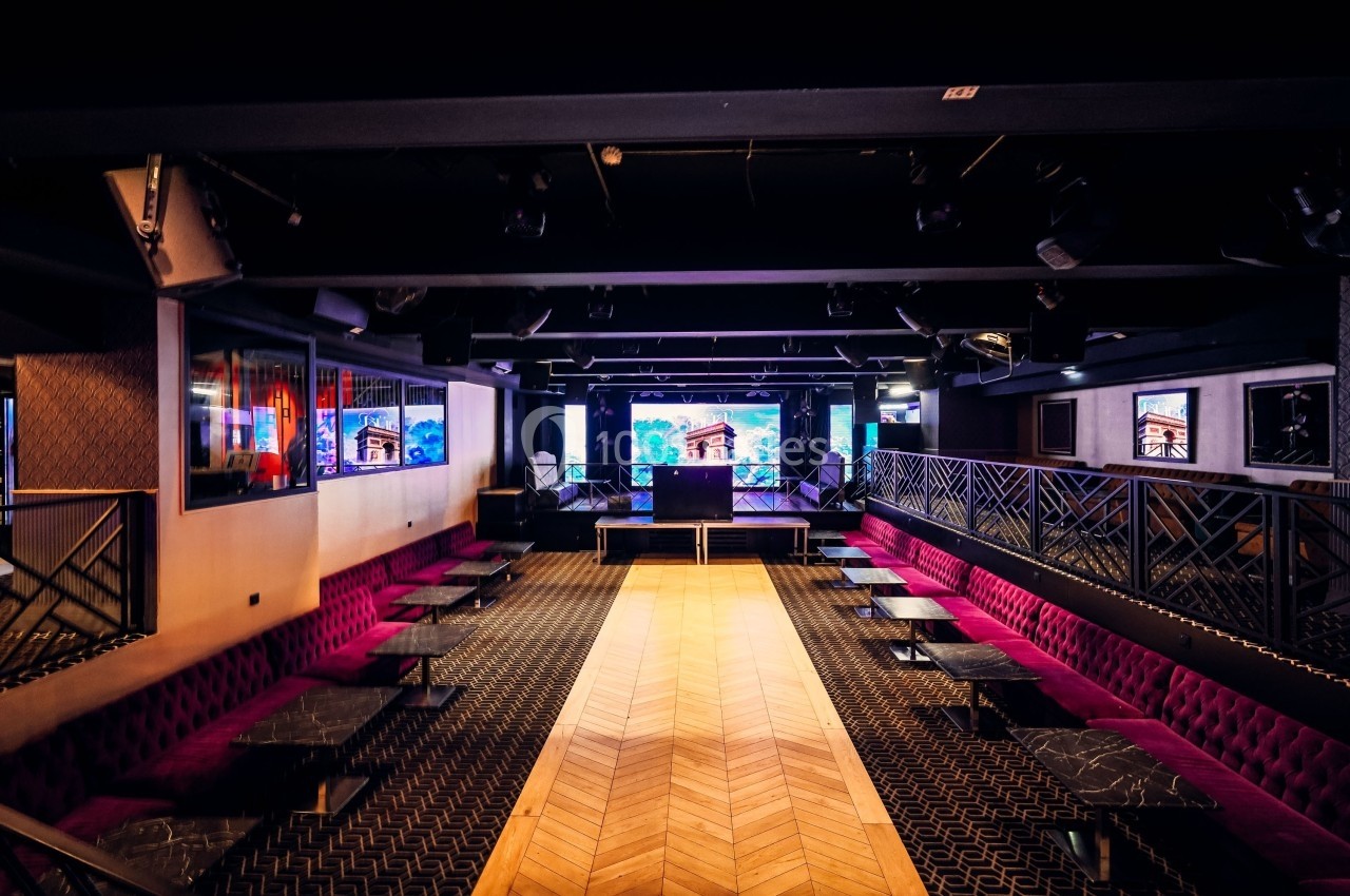 Salle de spectacle moderne avec scène centrale, banquettes violettes, tables noires et écrans muraux.