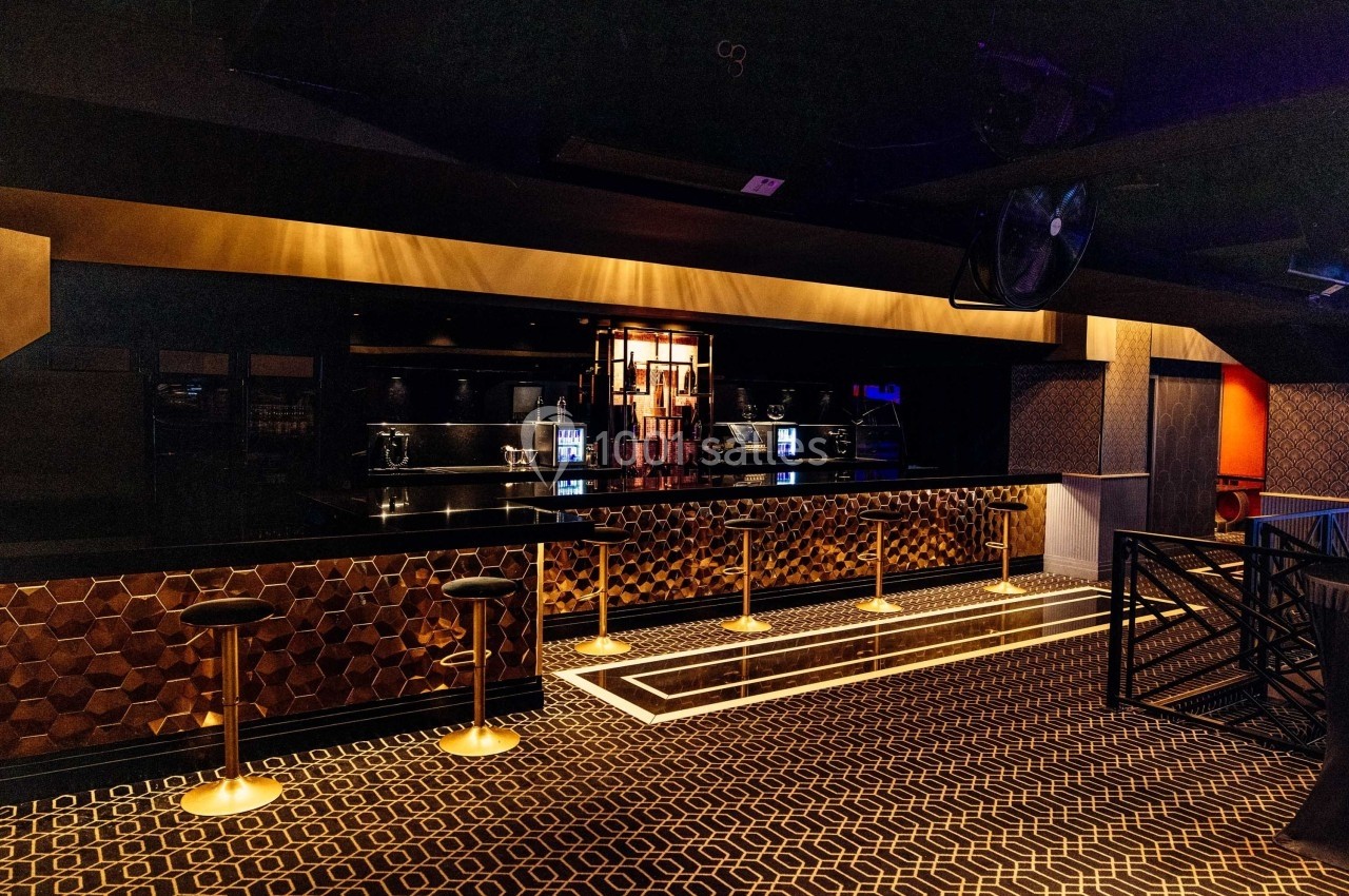 Intérieur d'un bar moderne avec comptoir en bois foncé, tabourets dorés et éclairage tamisé.