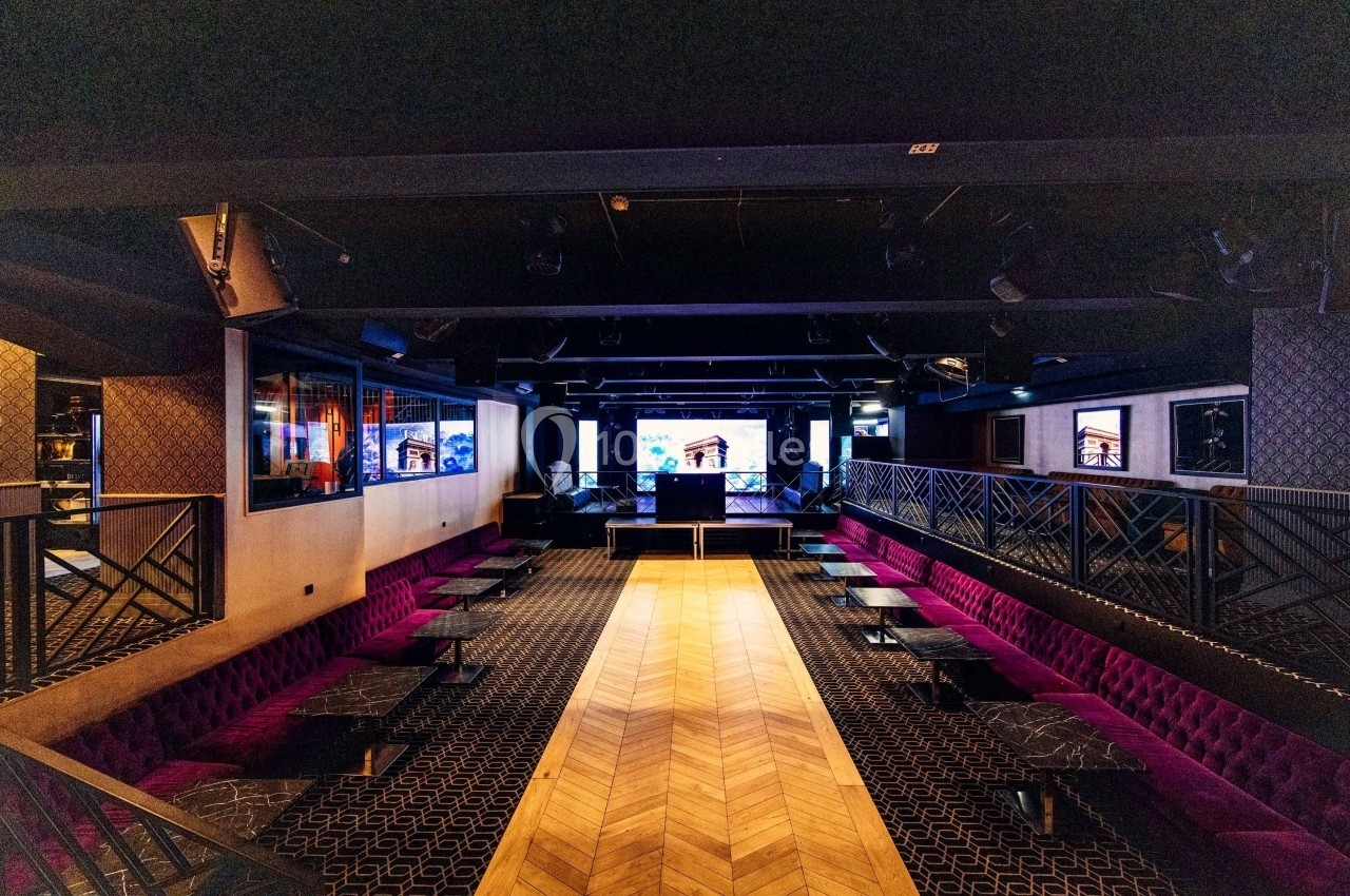 Salle de spectacle avec scène centrale, banquettes rouges, tables noires et éclairage tamisé.