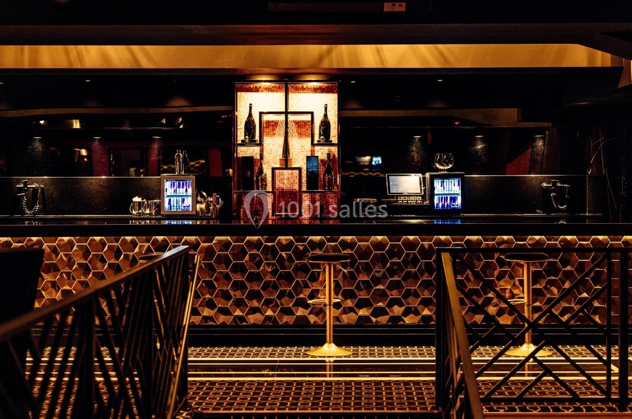 Bar élégant avec comptoir en mosaïque hexagonale, tabourets dorés et étagères illuminées exposant des bouteilles.