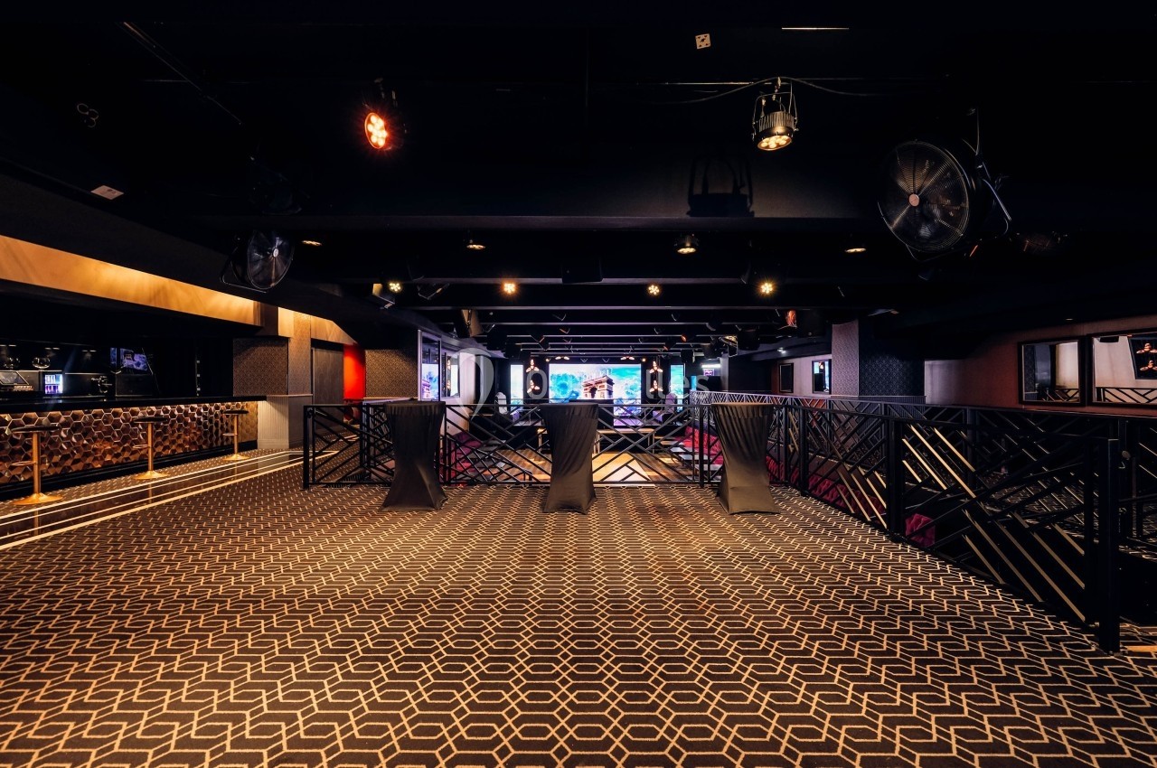 Salle sombre avec moquette géométrique, balustrades noires, éclairage tamisé et écran lumineux en arrière-plan.