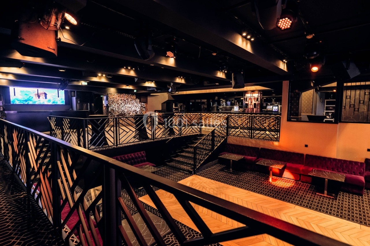 Salle moderne avec mezzanine, éclairage tamisé, banquettes rouges et bar en arrière-plan.