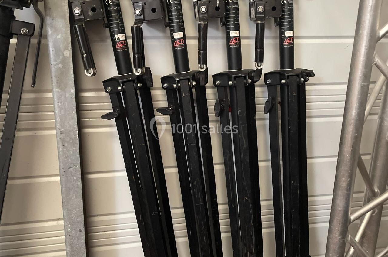 Quatre trépieds noirs pliés alignés verticalement contre un mur blanc dans un espace de stockage.