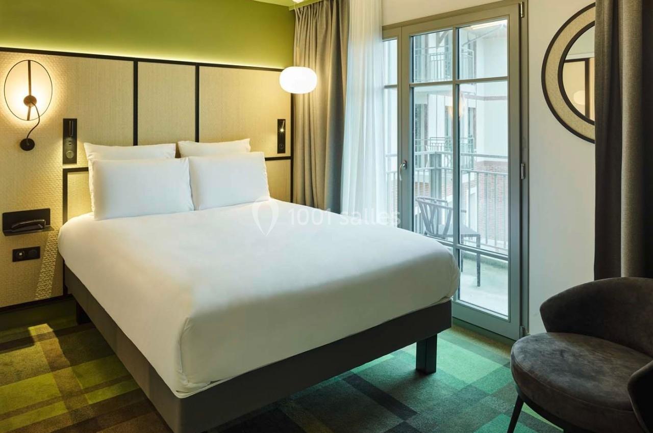 Chambre d'hôtel moderne avec lit double, éclairage mural, fauteuil, et accès à un balcon par une porte vitrée.