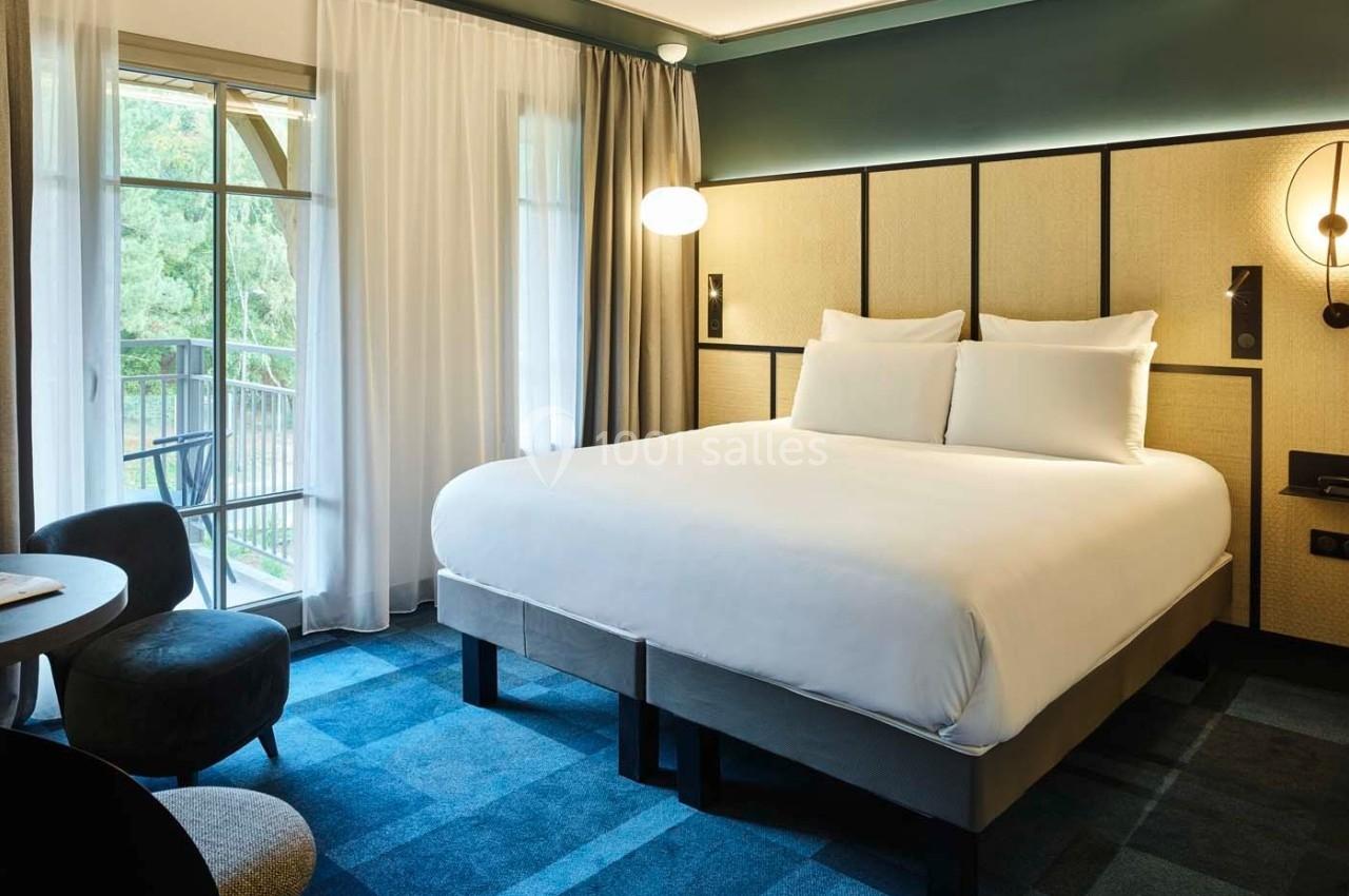 Chambre d'hôtel moderne avec un grand lit, un bureau, des fauteuils et une baie vitrée donnant sur un espace vert.