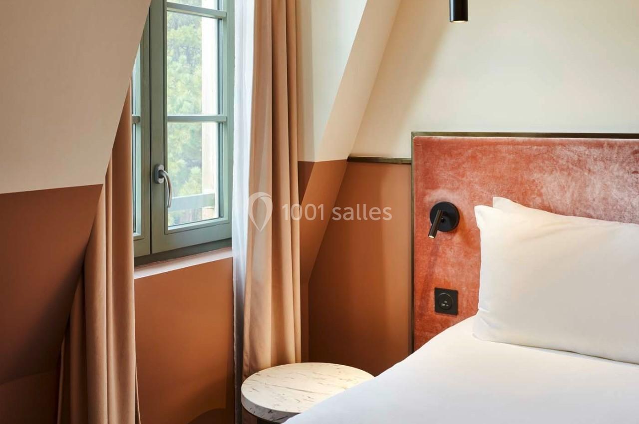 Lit avec tête de lit en velours, table d'appoint ronde et fenêtre avec rideaux dans une chambre lumineuse.