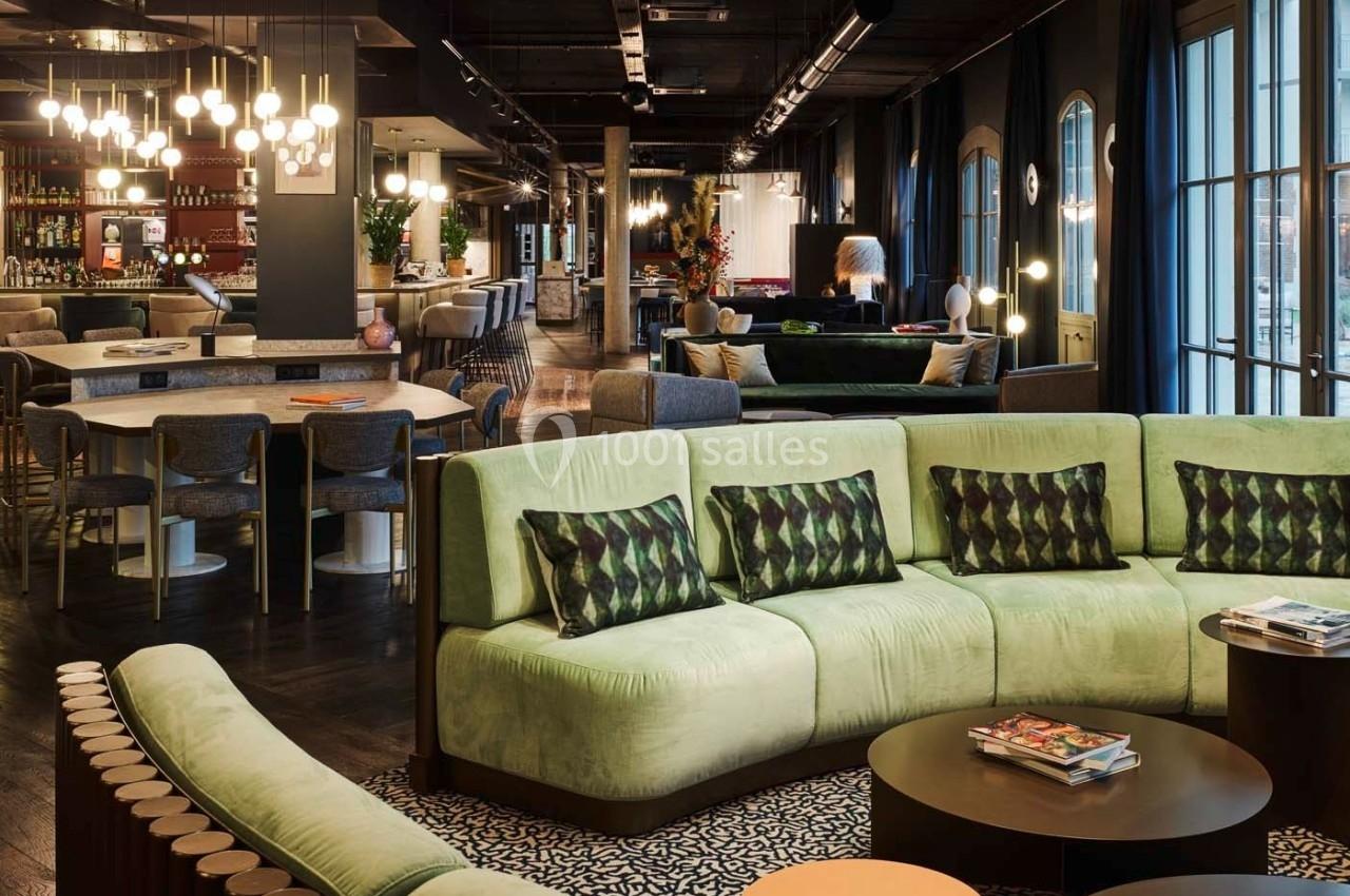 Salon moderne avec canapés verts, tables basses, bar éclairé et grandes fenêtres laissant entrer la lumière naturelle.