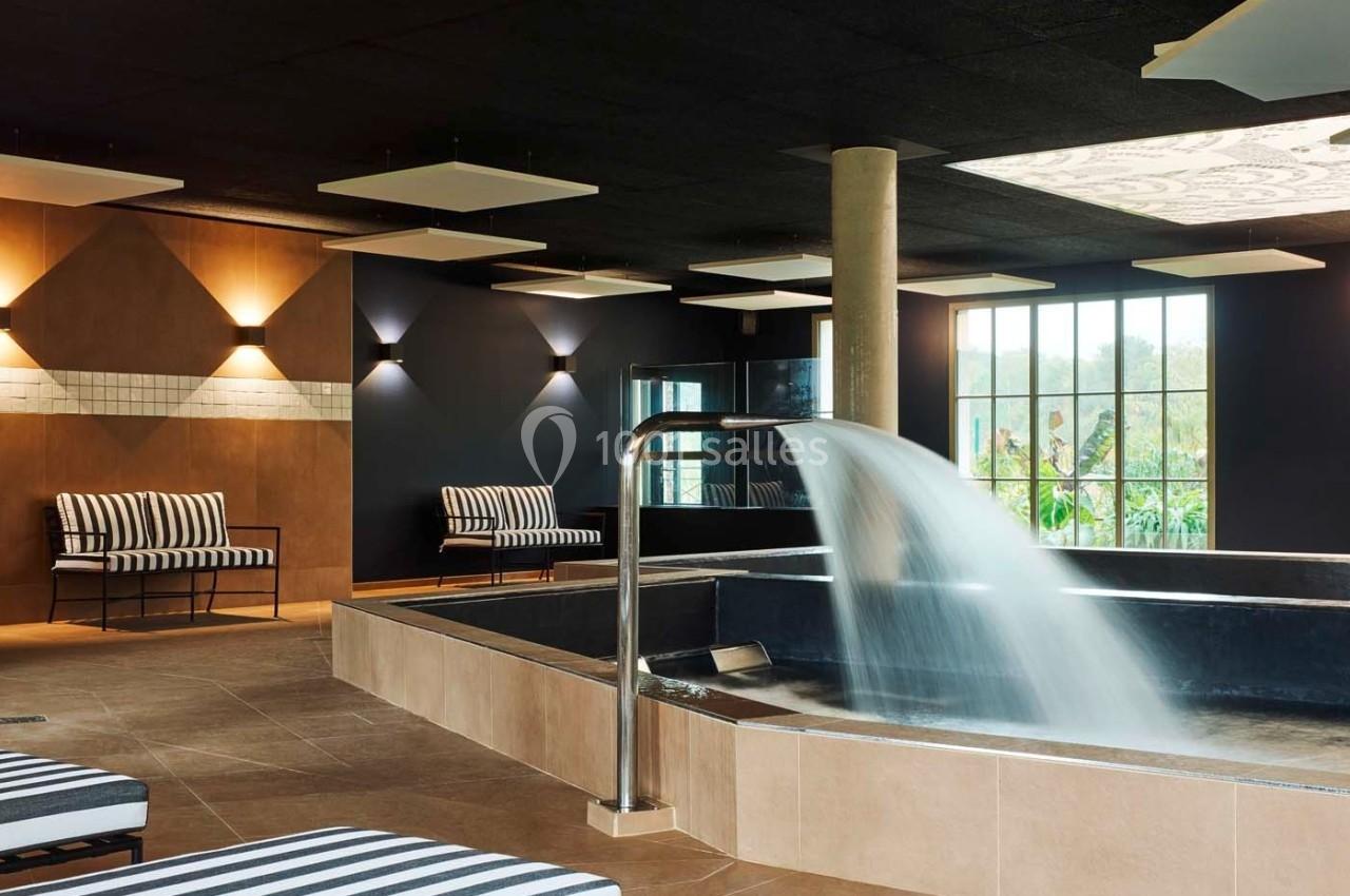 Salle de spa avec bassin, cascade d'eau, chaises longues rayées et grandes fenêtres donnant sur un jardin.